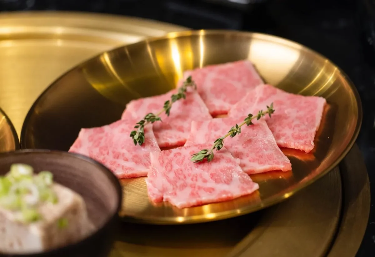 Plato dorado con finas lonchas de carne de res, acompañadas de un pequeño cuenco con tofu y cebollín, ideal para una comida gourmet.