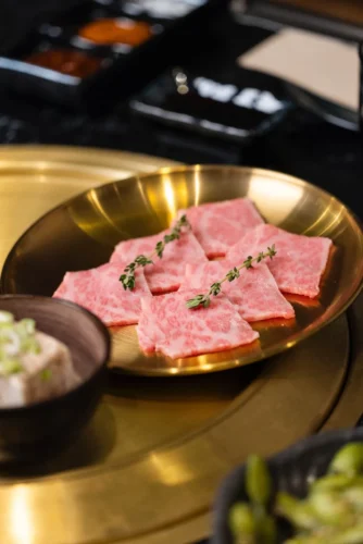 Plato dorado con finas lonchas de carne de res, acompañadas de un pequeño cuenco con tofu y cebollín, ideal para una comida gourmet.