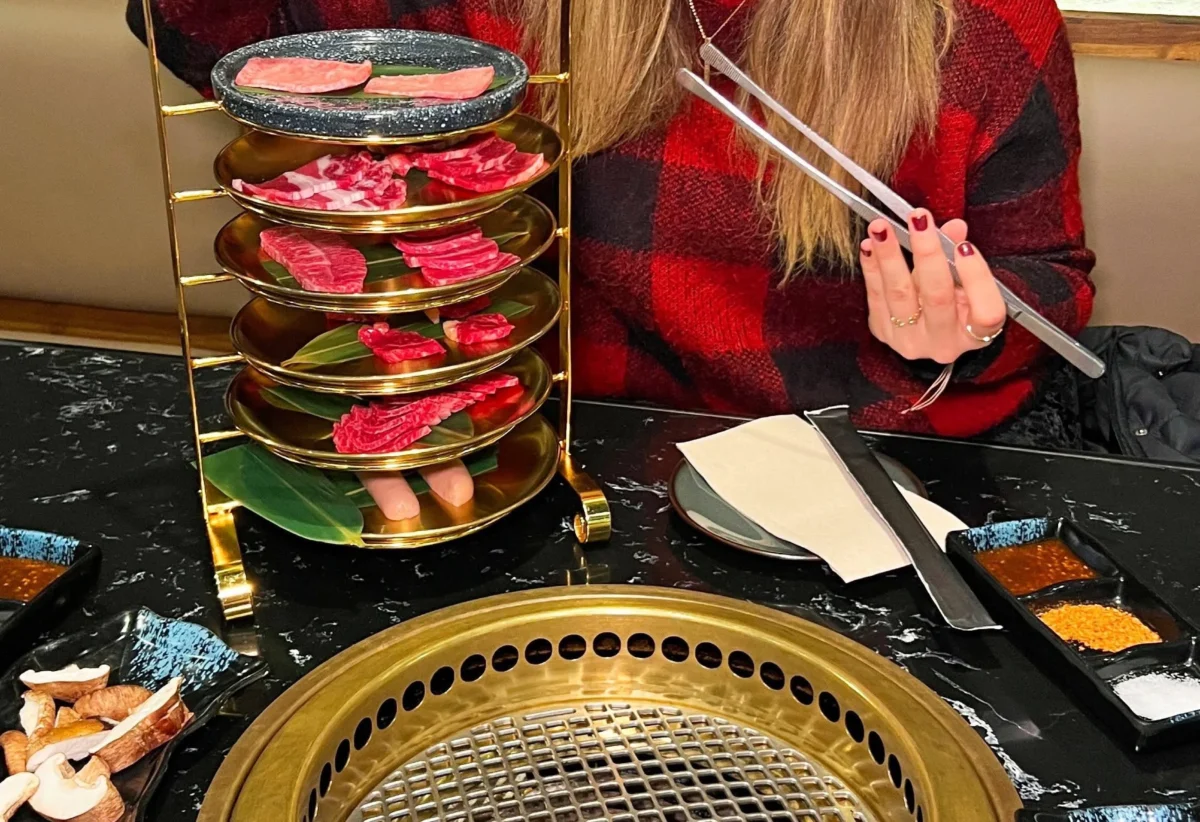 Mujer sonriente sosteniendo palillos frente a una parrilla de barbacoa, con platos de carne y vegetales en una mesa de restaurante.