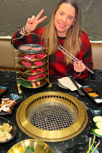 Mujer sonriente sosteniendo palillos frente a una parrilla de barbacoa, con platos de carne y vegetales en una mesa de restaurante.
